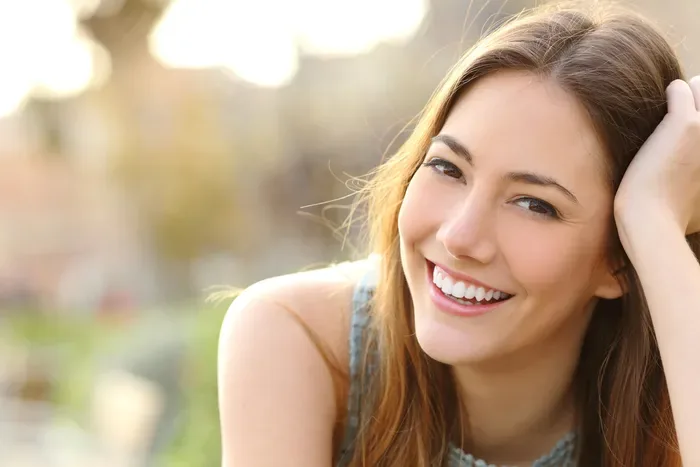 A smiling person outdoors resting a hand near their head, showing their teeth.